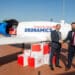 Two men shake hands in front of a cargo drone with parcels stacked next to them.